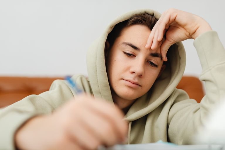Teenage boy in hoodie writing homework at home with headache.